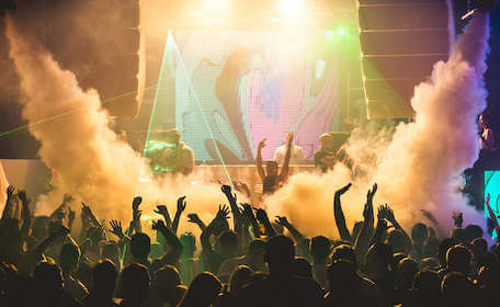 DJ on stage playing to large dancing crowd with lights and smoke machines